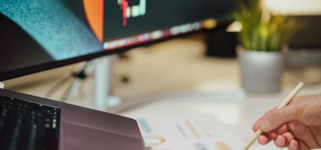 a person writing on a piece of paper next to a computer monitor by Jakub Żerdzicki courtesy of Unsplash.